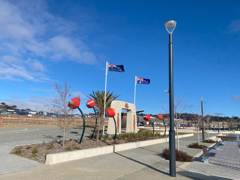 An ANZAC memorial area with large red poppy sculptures and two Australian flags flying above a stone commemorative arch, set against a bright blue sky. Modern pathway lighting lines the paved walkway beside the landscaped space.